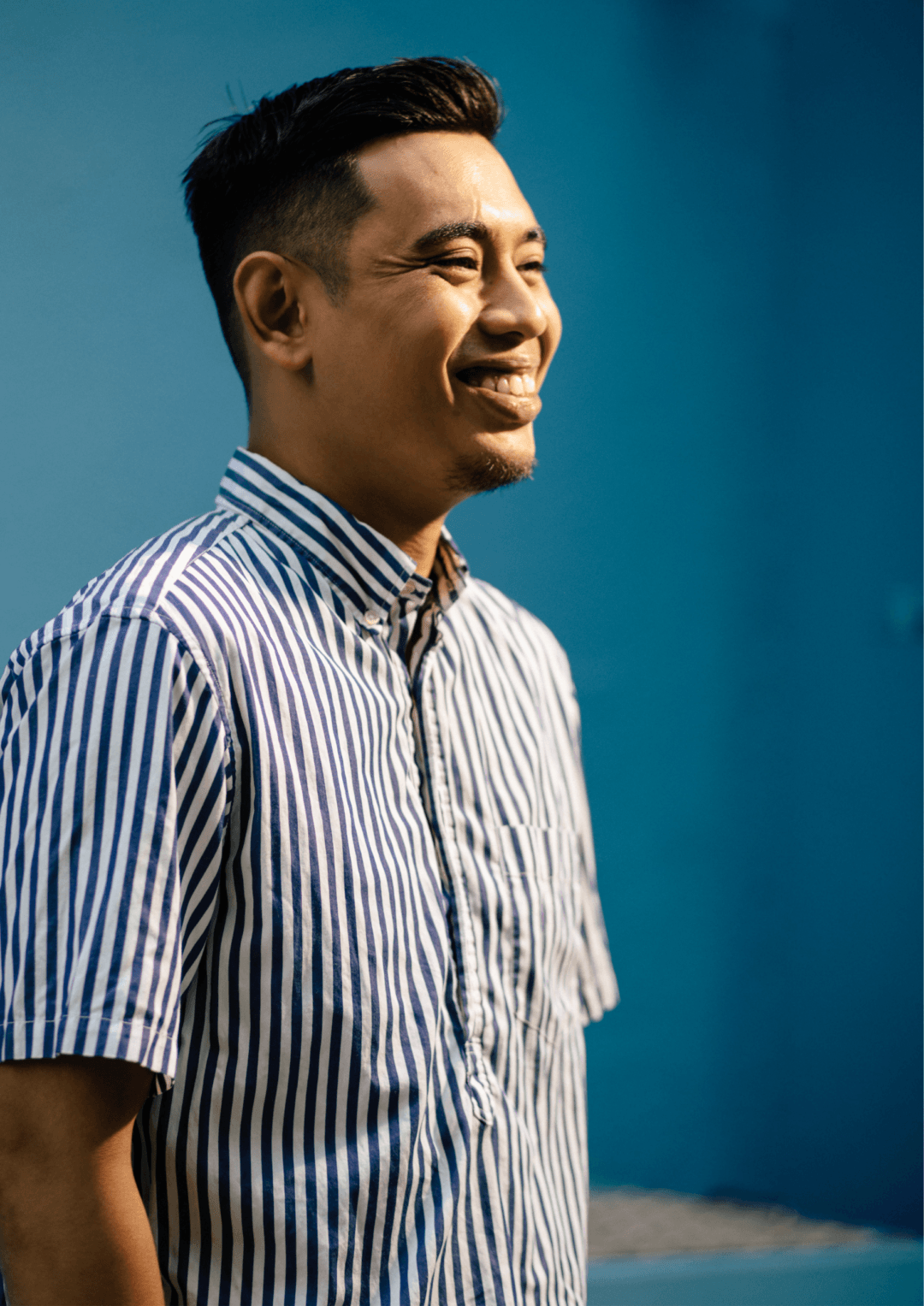 A side profile of a Kevin. He is standing against a cerulean blue wall, in a blue and white striped shirt. He is smiling.