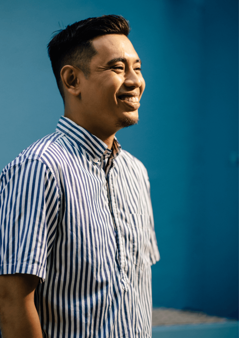A side profile of a Kevin. He is standing against a cerulean blue wall, in a blue and white striped shirt. He is smiling.